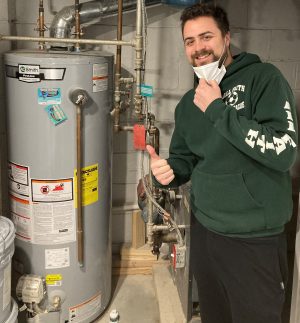 A person in a green hoodie stands next to a water heater in a basement, giving a thumbs up and holding a face mask under their chin.