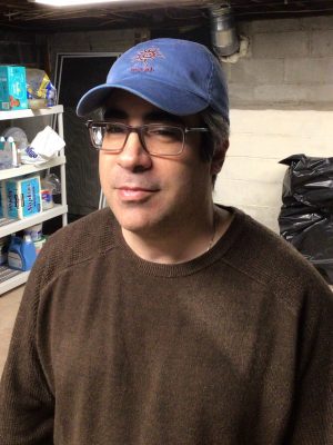 A man wearing glasses and a blue baseball cap stands in a basement with shelves of household items and a garbage bag in the background.