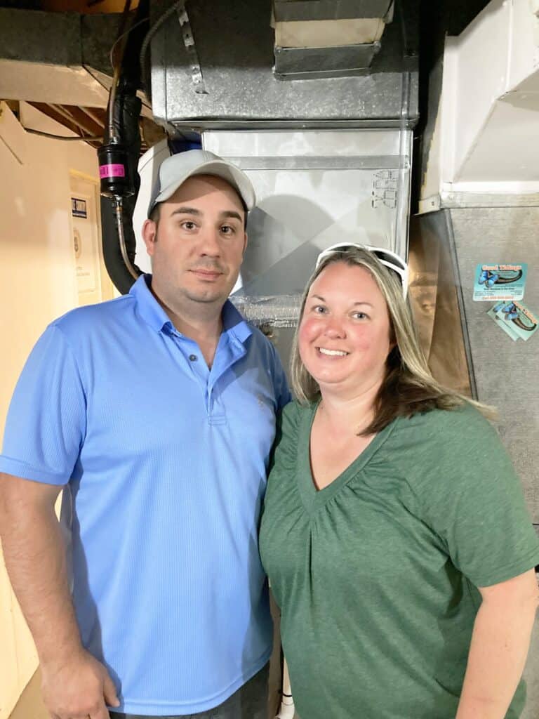 A man in a blue polo shirt and cap stands next to a woman in a green shirt, both indoors near metal HVAC ducts.