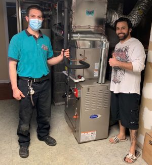 Two men stand next to a newly installed furnace; one is wearing a mask and uniform, the other is smiling and giving a thumbs up.