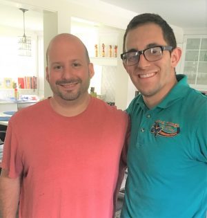 Two men are standing indoors, smiling at the camera. One is wearing a red shirt, and the other is wearing glasses and a teal polo shirt. The background shows a bright kitchen area.
