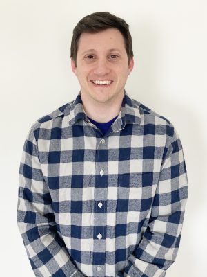 A man with short brown hair wearing a blue and white plaid shirt stands in front of a plain white background, smiling at the camera.