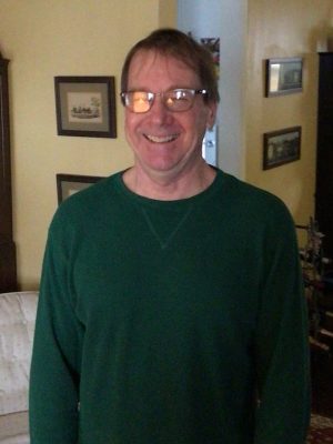 A man wearing glasses and a green sweatshirt stands indoors, smiling at the camera. Framed pictures decorate the yellow wall behind him.