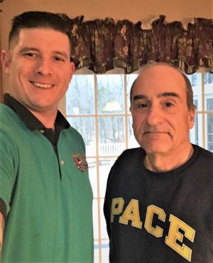 Two men stand indoors, smiling at the camera. One wears a green polo shirt, and the other wears a dark sweatshirt with the word "PACE" in yellow letters. A window is in the background.