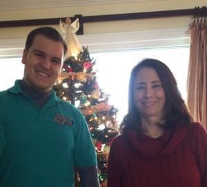 Two people stand and smile in front of a decorated Christmas tree inside a room with a window and curtains.