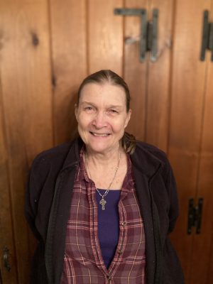 An older woman with long hair smiles at the camera, standing in front of wooden paneled doors. She wears a plaid shirt, dark jacket, and a necklace.