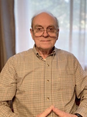 Older man with glasses and a checked shirt sits indoors in front of a window with sheer curtains, hands resting together in front of him.