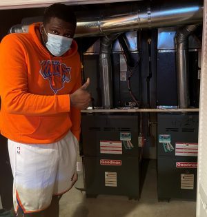 A person in an orange hoodie and mask gives a thumbs up while standing in front of two Goodman HVAC units in a utility area.