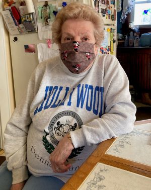 An elderly woman wearing a face mask and a gray "Hollywood, Pennsylvania" sweatshirt sits at a kitchen table.