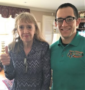 An older woman gives a thumbs up while standing next to a younger man in glasses and a green polo shirt. They are indoors with sunlight coming through a window.