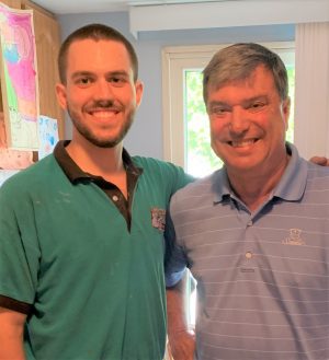 Two men standing side by side indoors, smiling at the camera. There is a window and children's artwork visible in the background.