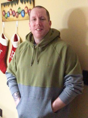 A man in a green and gray hoodie stands indoors with his hands in his pockets; two Christmas stockings hang on the wall behind him.