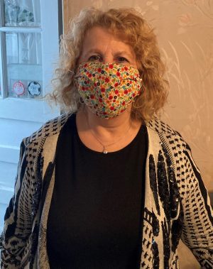 A woman with curly hair wears a colorful patterned face mask, a black top, and a patterned cardigan, standing indoors near a window.