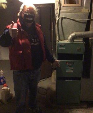 Person wearing a mask and red vest stands in a dimly lit basement, giving a thumbs up while pointing at a green furnace next to ventilation pipes.