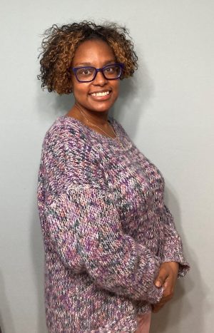 A woman with curly hair wearing glasses and a multicolored knit sweater stands against a plain light gray background, smiling at the camera.