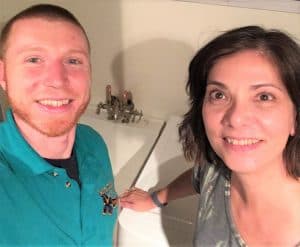 Two people standing and smiling for a selfie in front of a white bathtub with silver fixtures.