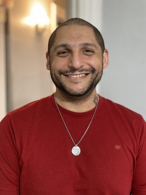 A man with short hair and a beard smiles at the camera. He is wearing a red shirt and a necklace with a round pendant. The background is softly lit and out of focus.