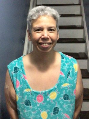 A woman with short gray hair smiles while standing in front of a staircase, wearing a turquoise top with a colorful turtle pattern.