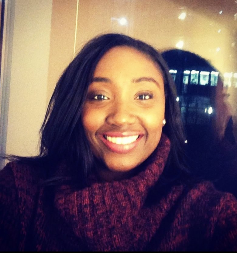 A woman with long dark hair and a maroon sweater smiles at the camera indoors with warm lighting in the background.