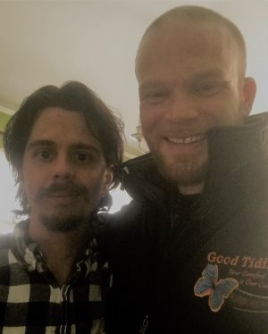 Two men pose for a photo indoors; one wears a plaid shirt, the other a black jacket with a butterfly logo and the words “Good Tid.”.