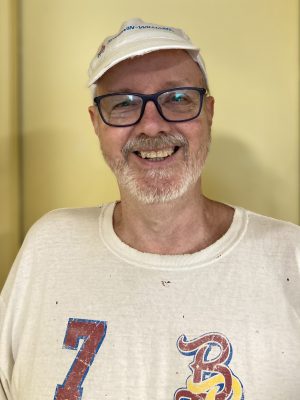 A man with glasses and a white cap smiles at the camera, wearing a white T-shirt with colorful numbers and letters printed on it.