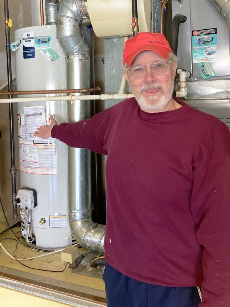 An older man in a red cap and maroon sweater stands in a utility room, pointing at a water heater beside other household equipment.
