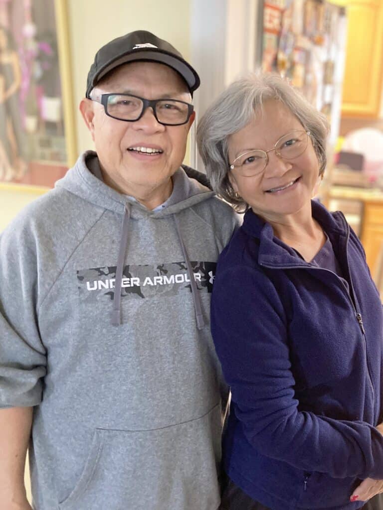 Two older adults, one wearing glasses and a gray hoodie, and the other with glasses and a dark jacket, stand close together and smile indoors.