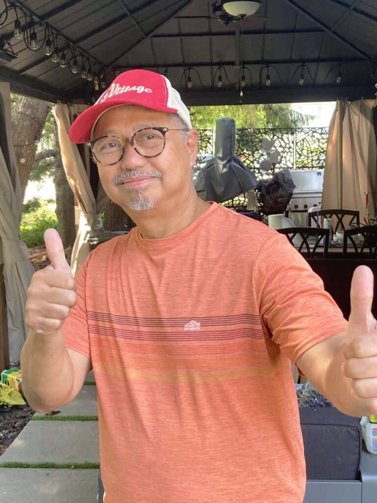 An older man wearing glasses, a red cap, and an orange shirt stands under a gazebo giving two thumbs up.