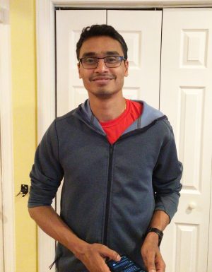 A man wearing glasses and a blue hoodie stands indoors in front of white double doors, holding a smartphone.