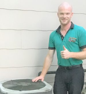 A person standing outside next to an air conditioning unit, giving a thumbs up, and wearing a green collared shirt and black pants.