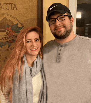 A woman with long red hair and a scarf stands next to a man wearing glasses, a black cap, and a gray shirt. They are both smiling indoors.