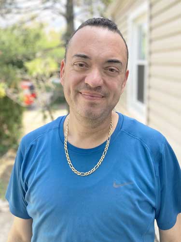 A man wearing a blue Nike t-shirt and a gold chain stands outside near a beige house, smiling at the camera.