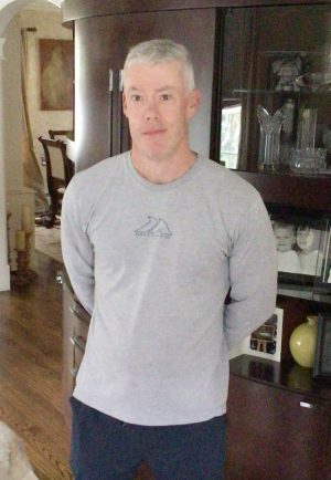 A man with short gray hair wearing a light gray long-sleeve shirt and dark pants stands indoors in front of a dark wooden cabinet.