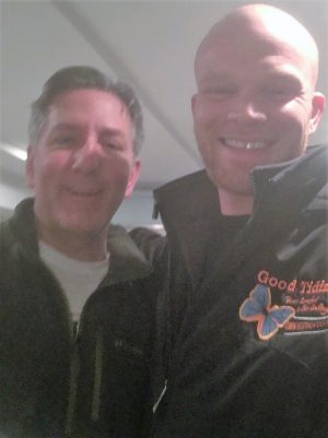 Two men are standing close together indoors, both smiling at the camera. The man on the right is wearing a jacket with an embroidered logo and butterfly design.