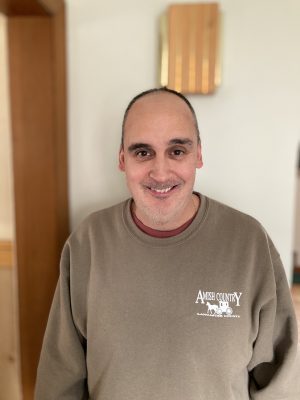 A man with a receding hairline is standing indoors, wearing a brown "Amish Country" sweatshirt and smiling at the camera.