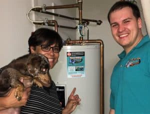 A woman holding a dog and a man stand next to a water heater; the woman points at a sticker on the heater and both are smiling at the camera.