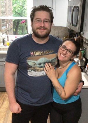 A man and woman stand smiling together in a kitchen, with the woman resting her hand on the man's chest. Both are wearing glasses and casual clothes.