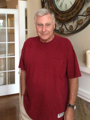 An older man with gray hair stands indoors wearing a red t-shirt and light-colored pants, with one hand in his pocket and a large wall clock behind him.