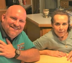 A man and an older woman sitting together at a kitchen table, both looking at the camera and smiling.