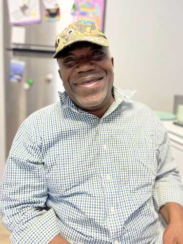 A man wearing a camouflage cap and a checked shirt sits indoors, smiling at the camera. A refrigerator and wall decorations are in the background.