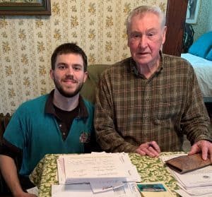 Two men sit at a table covered with papers, a notebook, and a pen. The younger man smiles at the camera while the older man looks forward. A bedroom is visible in the background.