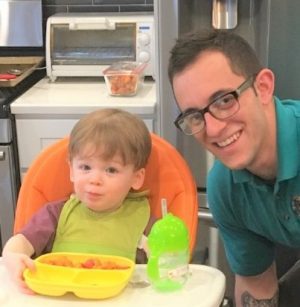 A man wearing glasses smiles next to a young child sitting in a high chair with food and a green sippy cup on the tray in a kitchen setting.