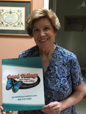 A woman in a blue patterned shirt holds a Good Tidings Plumbing Heating Cooling folder, standing indoors and smiling at the camera.
