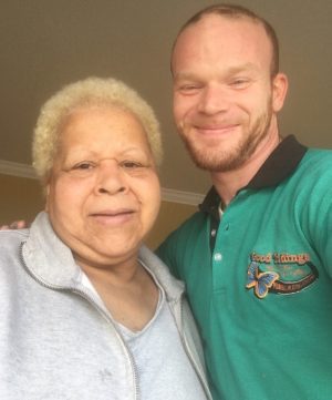 Two people stand close together indoors, smiling at the camera. The person on the left has light hair and wears a gray zip-up, while the person on the right wears a green collared shirt.