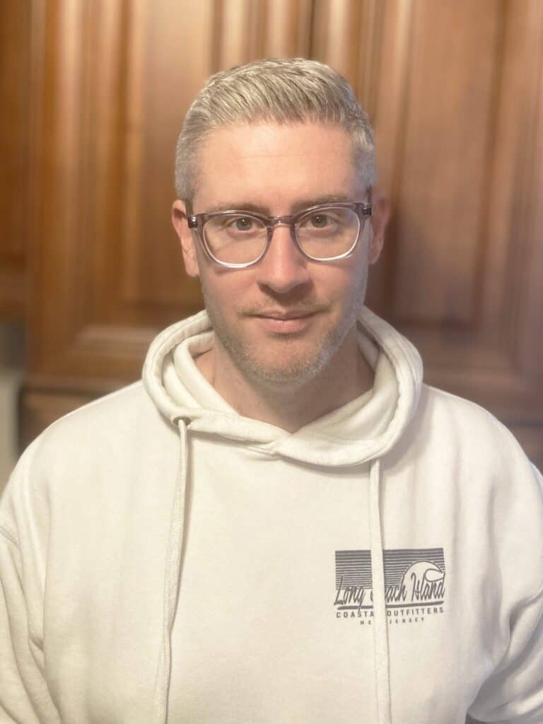 A man with short, light hair and glasses is wearing a white hoodie with a graphic on the chest, standing indoors in front of wooden cabinets.