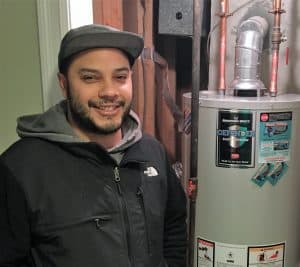 A man in a black jacket and cap stands next to a water heater in a utility area with exposed pipes and wooden beams.