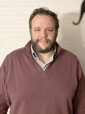 A man with light skin, a beard, and short hair wearing a maroon quarter-zip sweater over a checkered shirt stands in front of a light-colored wall.