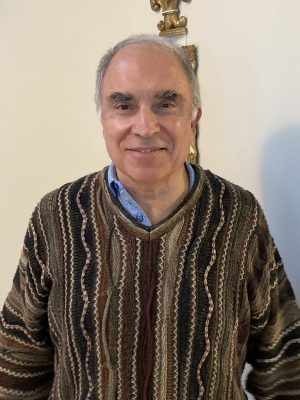 An older man with short gray hair wearing a patterned sweater over a blue shirt stands indoors, smiling at the camera.