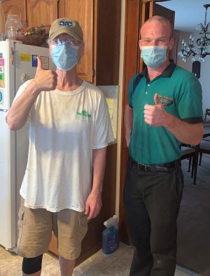 Two men wearing face masks stand indoors, giving thumbs up. One wears a tan hat and t-shirt; the other, a green polo and black pants. They are in a kitchen area.