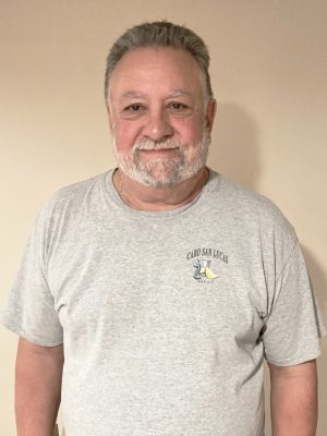 A man with gray hair and a beard is standing against a plain beige wall, wearing a light gray t-shirt with a small graphic on the left chest.